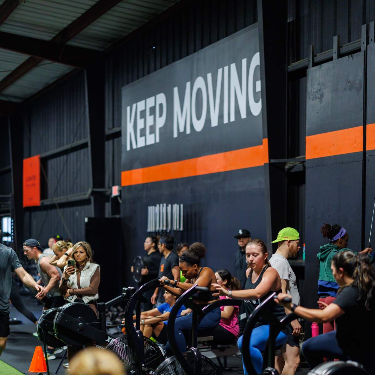 A group of people is engaged in a workout session at a fitness facility, using exercise bikes and rowing machines under a "KEEP MOVING" wall sign.