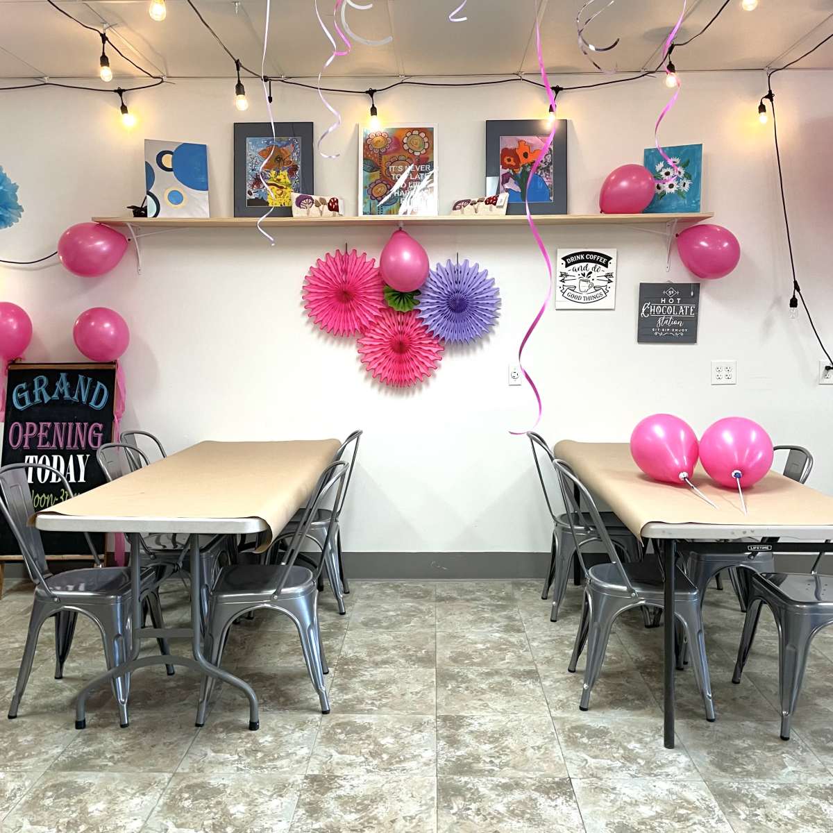 A decorated event space with two tables, pink balloons, and colorful paper flowers on the wall, set for a grand opening celebration.