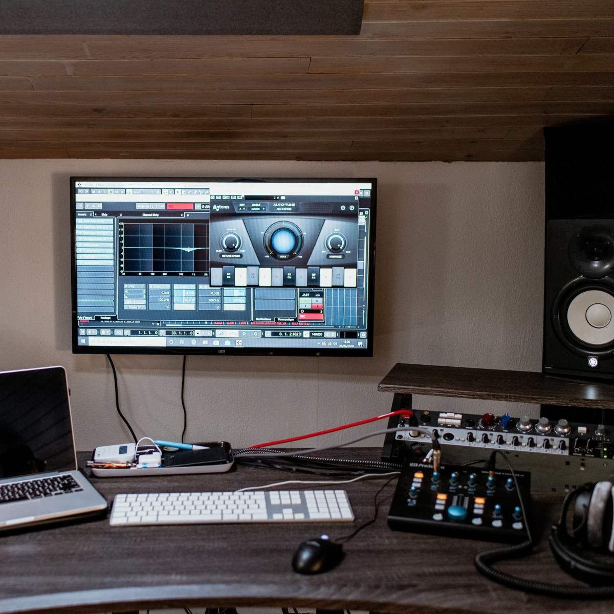 A music production workspace features a large monitor displaying software, flanked by two studio monitors, with a laptop and audio equipment on the desk.