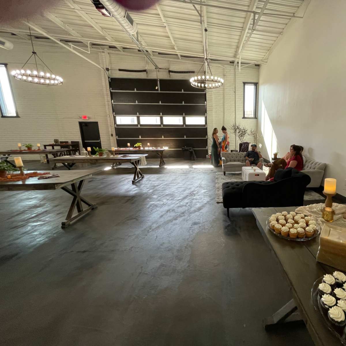 A spacious indoor venue with tables arranged for an event, featuring desserts on display, and several people seated in a lounge area.