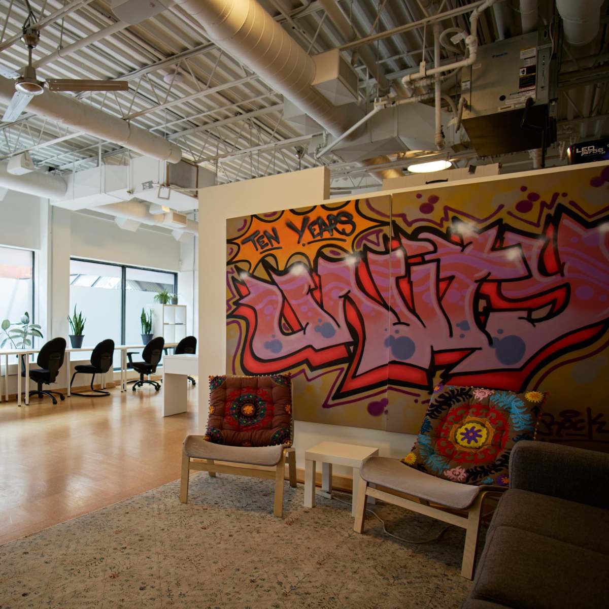 A modern workspace with a colorful graffiti wall, an area for seating, and several desks arranged in a well-lit room with plants and large windows.