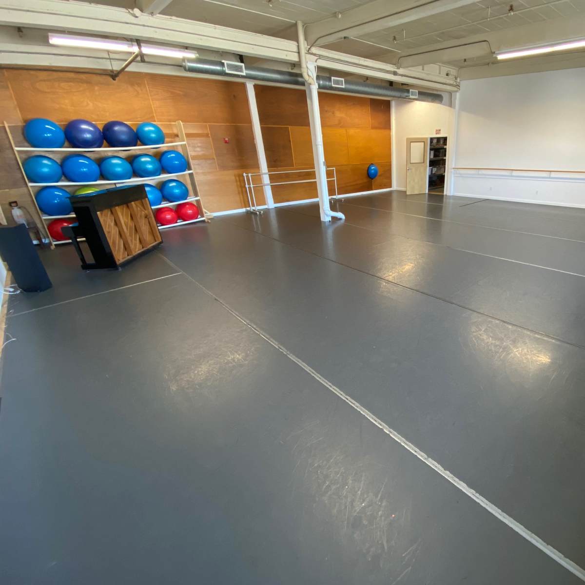 A spacious, empty exercise studio with gray flooring and blue exercise balls stacked against a wooden wall.