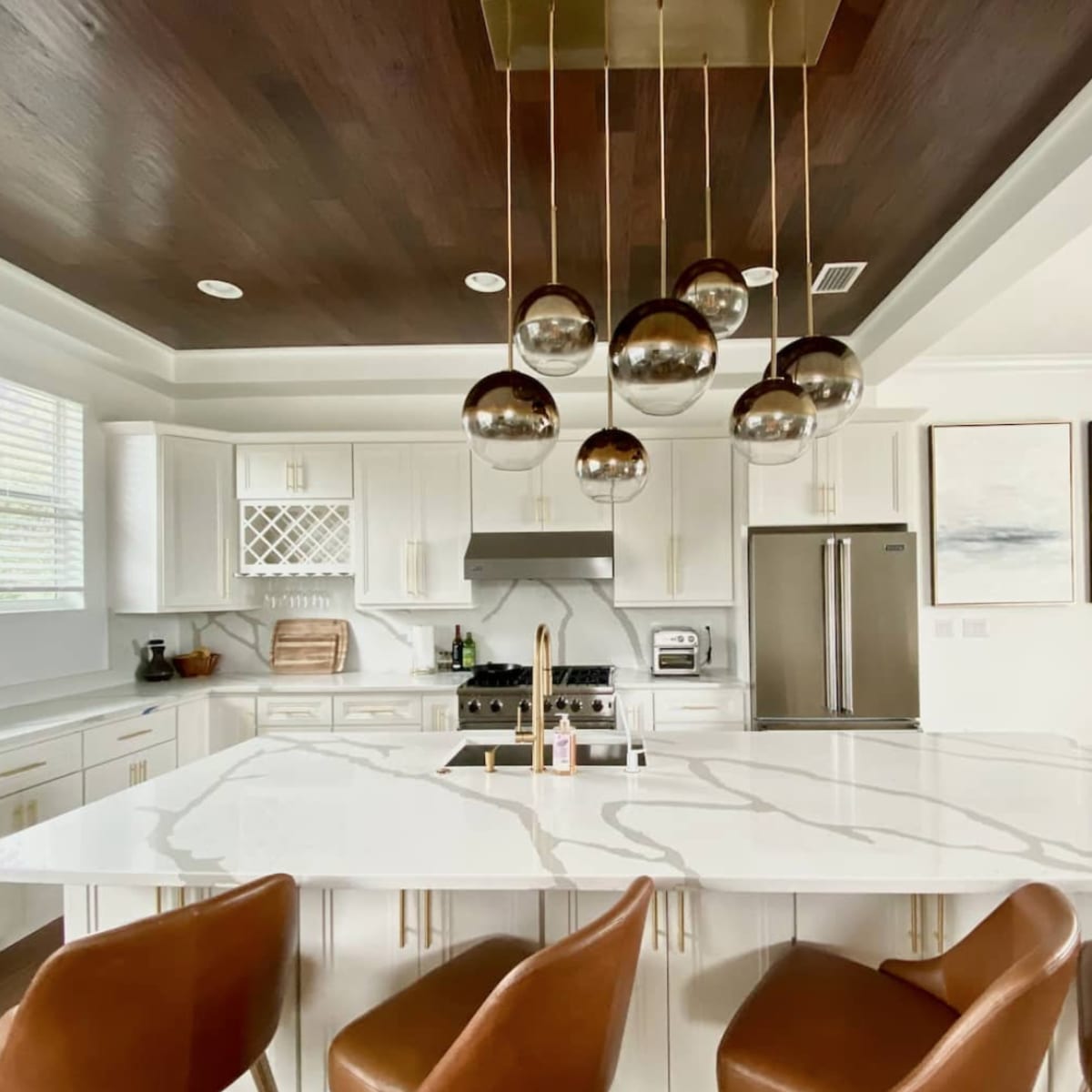 A modern kitchen features a large marble island with metal bar stools, pendant lighting, and stainless steel appliances.