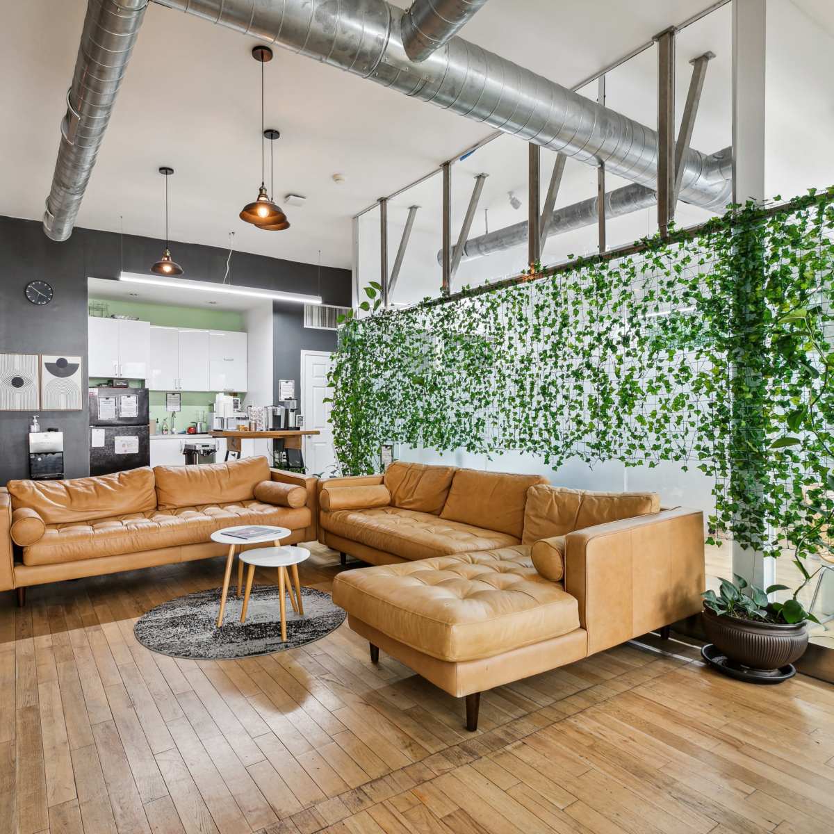 A modern living space featuring a brown leather sectional sofa, a small round table, and a wall adorned with greenery next to a kitchen area with appliances.