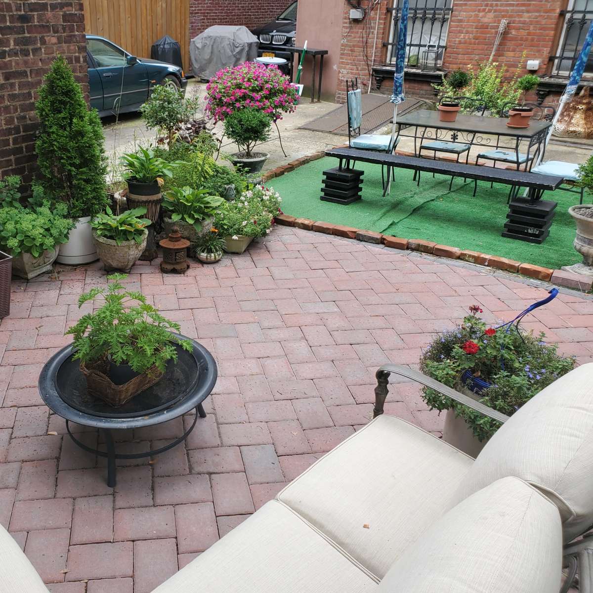 A brick-paved outdoor area features various potted plants, a small table, and seating arranged around a central fire pit.