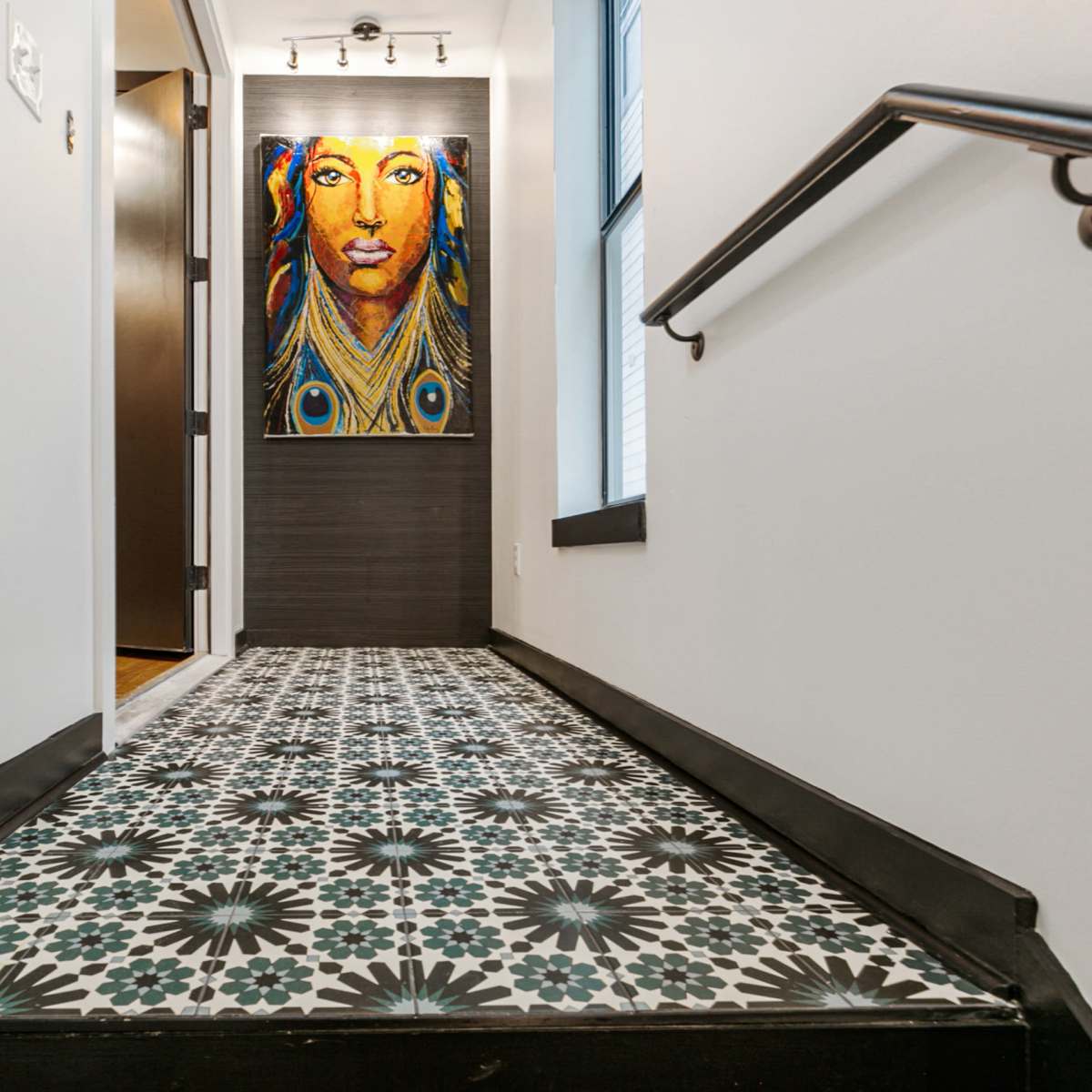 A hallway with patterned tile flooring leading to a dark door, illuminated by a brightly colored portrait on the wall.