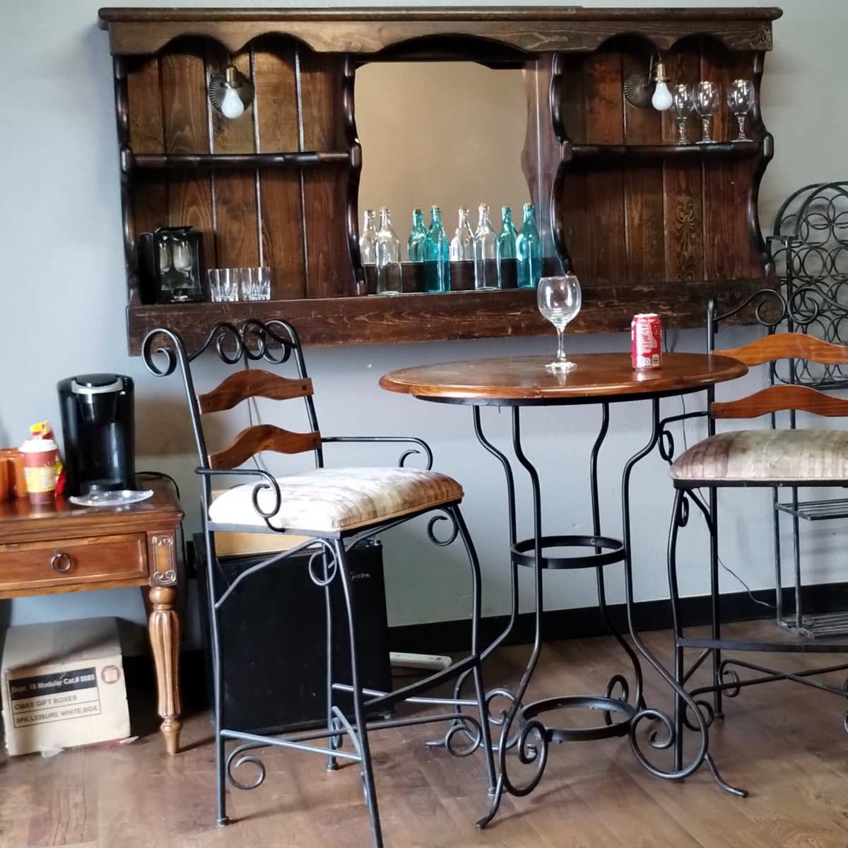 A small room with a wooden bar set featuring two metal stools, a mirrored cabinet, a table with a drink, and a neatly made bed in the background.