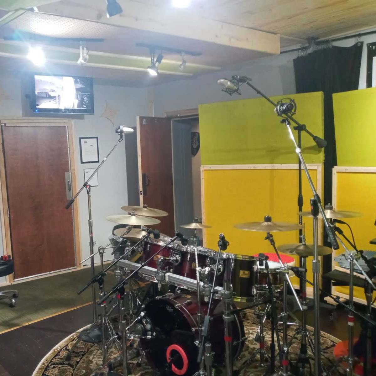 A music recording studio with a drum set, microphones positioned around it, and a television mounted on the wall.