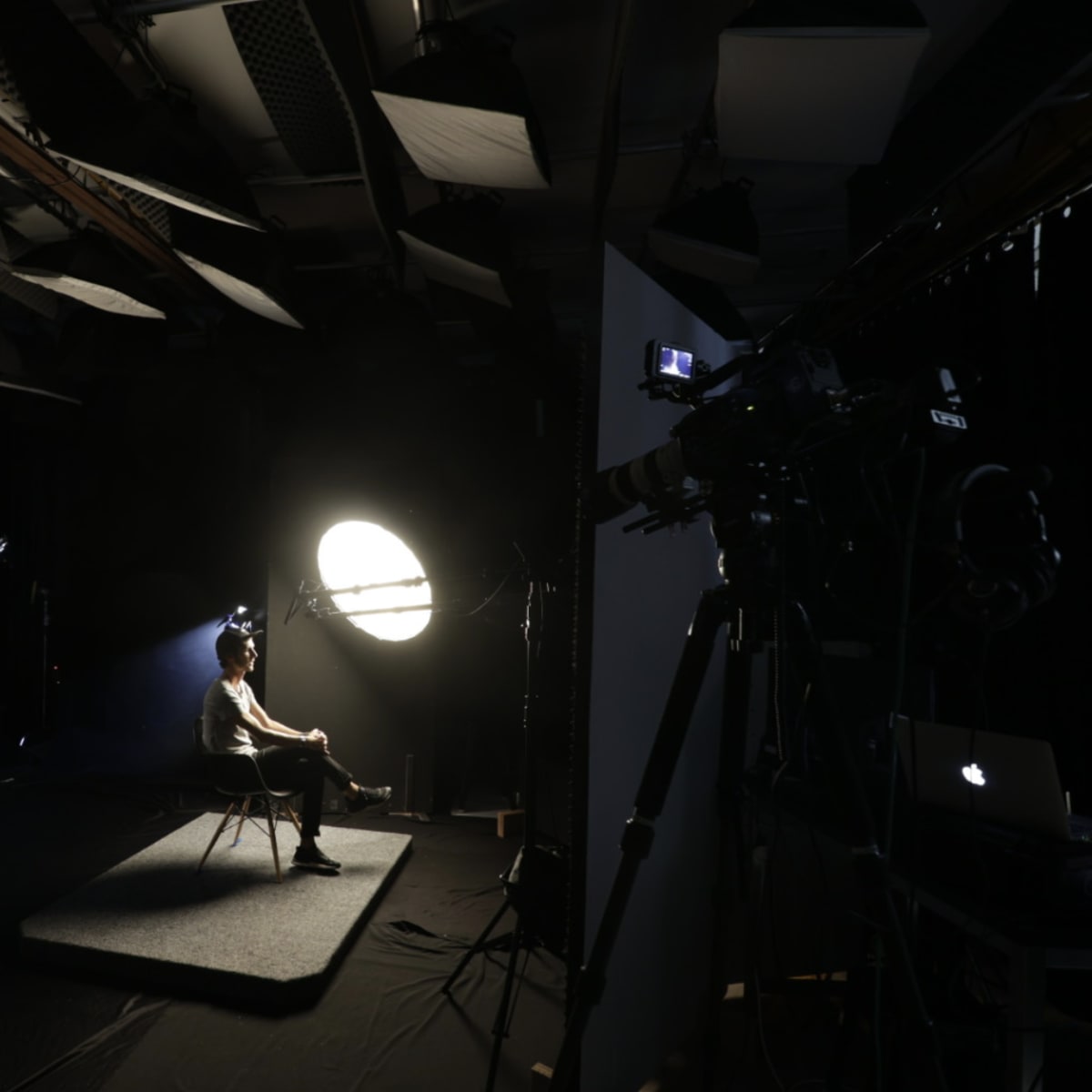 A person sits on a chair in a dimly lit studio with a large circular light illuminating them, surrounded by cameras and lighting equipment.