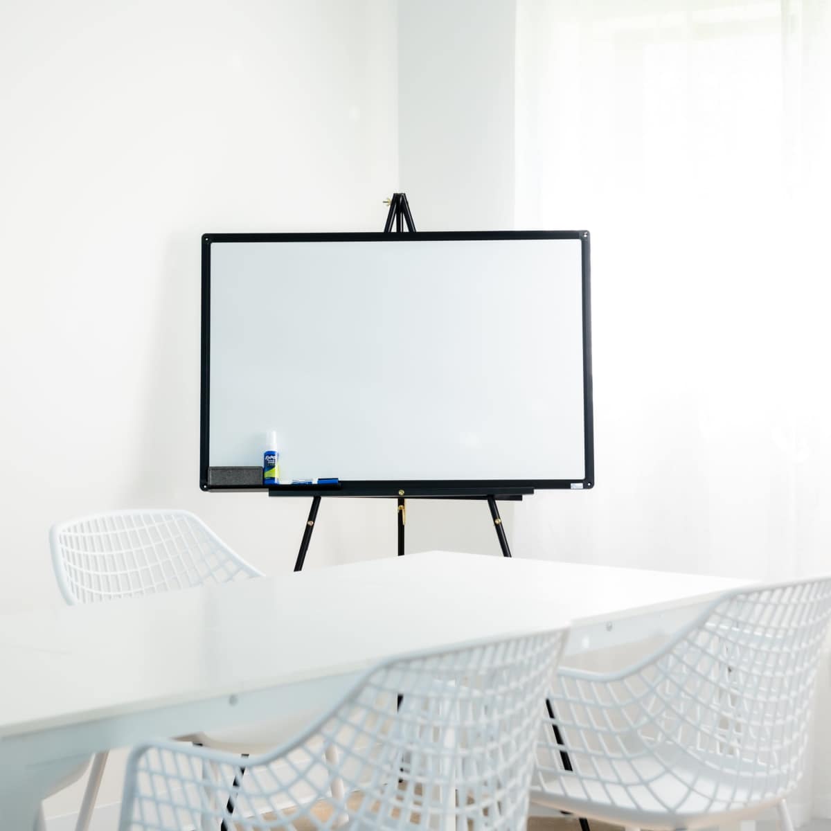 A whiteboard on an easel stands in a well-lit room with white walls and several white chairs around a table.