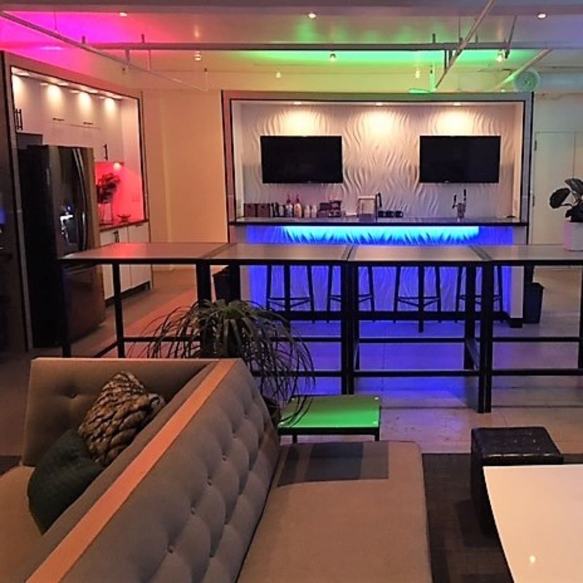 A modern lounge area with a bar featuring illuminated counters, two television screens, and a seating arrangement nearby.