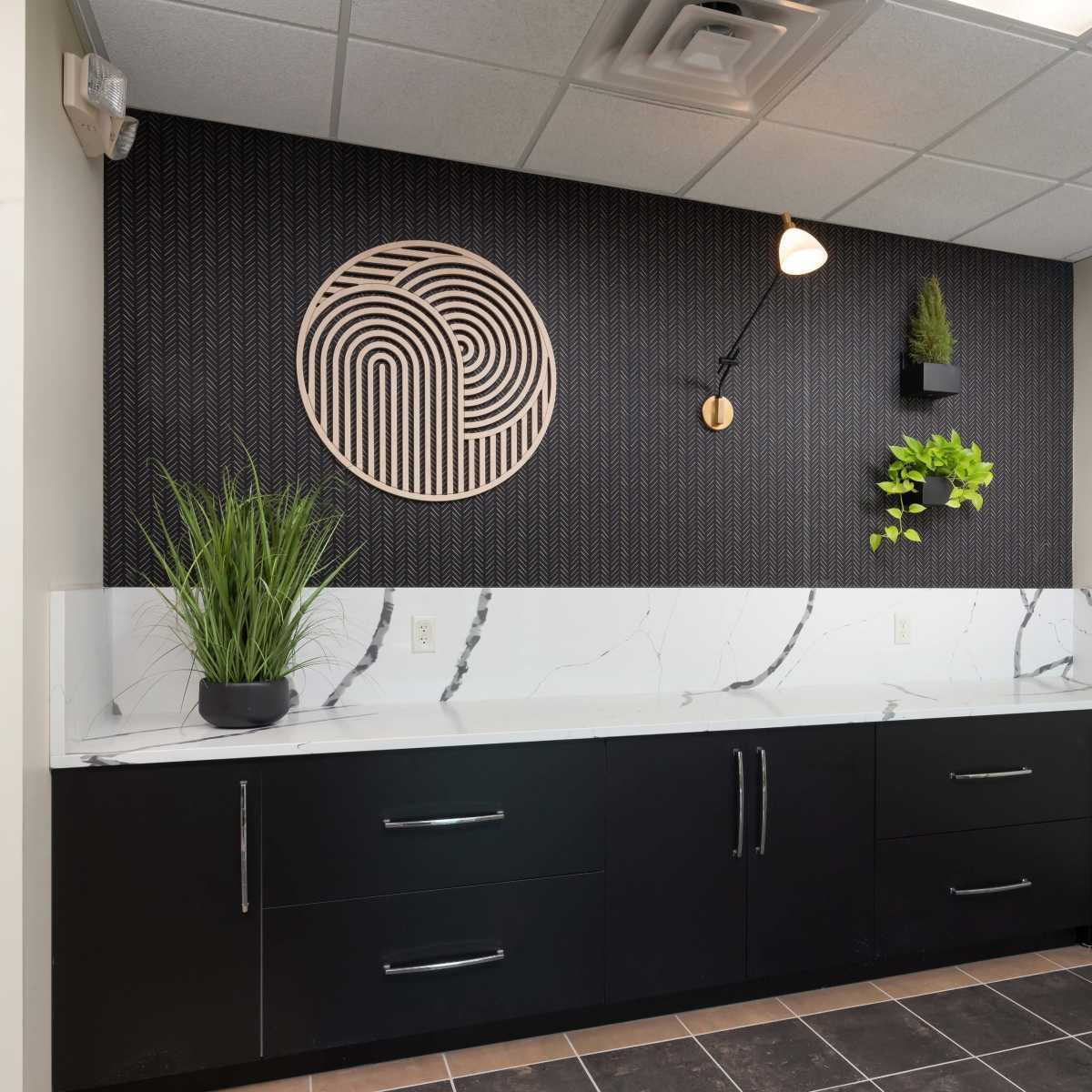 A modern kitchenette with a dark cabinet base, a white marble countertop, and a decorative wall featuring a circular design and greenery.