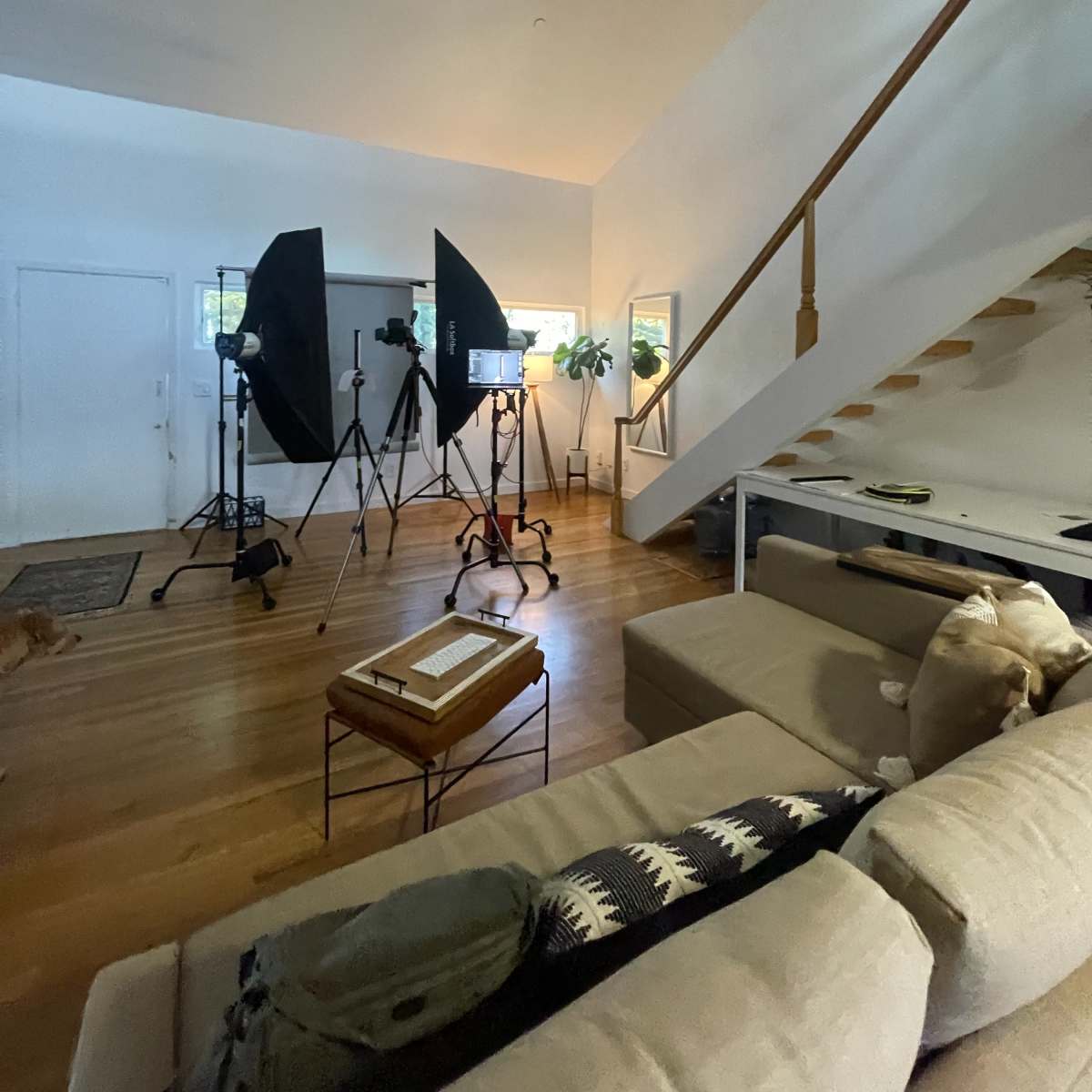 A brightly lit living room with video equipment set up, including tripods and softboxes, alongside a staircase and a sofa.
