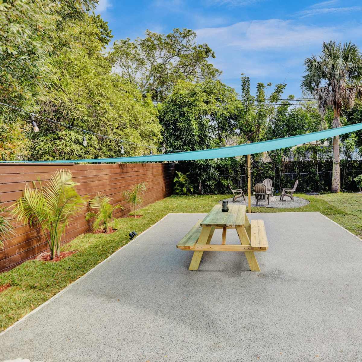A picnic table is positioned on a concrete patio surrounded by greenery and shaded by a blue fabric canopy.