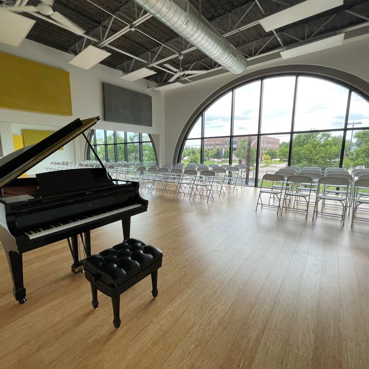 A grand piano is positioned near a large window in a spacious room filled with folding chairs set up for an event.