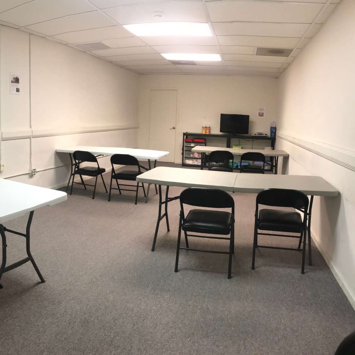A neatly arranged meeting room features several foldable tables and chairs set up for discussions, with a small desk and monitor in one corner.