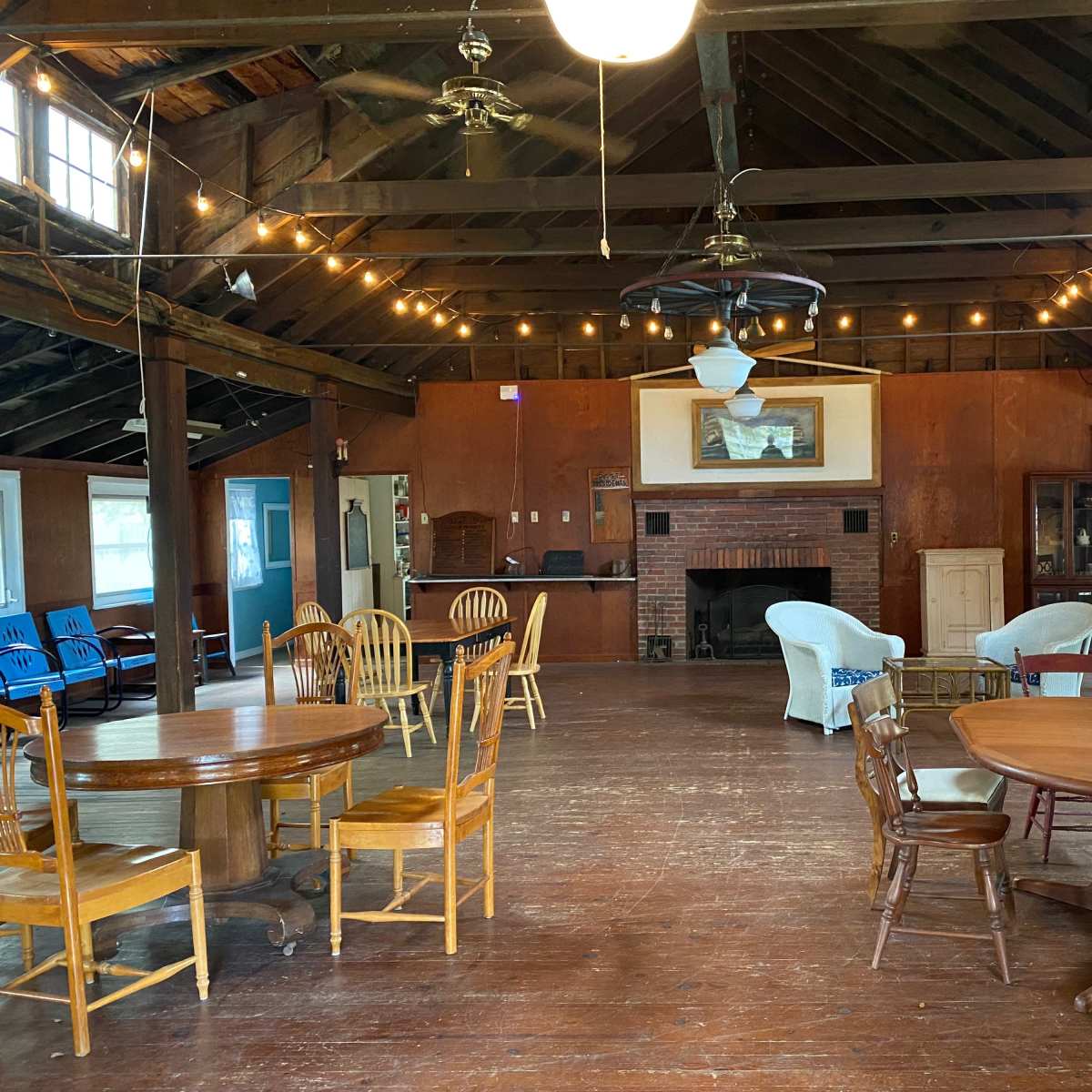 A spacious, wooden interior of a lodge with tables and chairs arranged in the open area, a fireplace in the back, and string lights hanging from the ceiling.