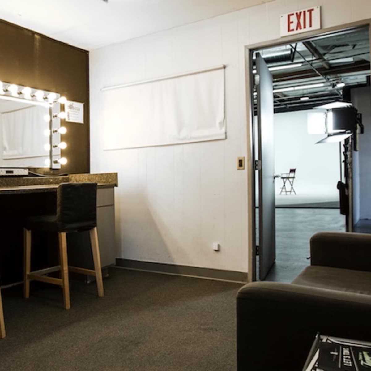 A dressing room with a large lighted mirror, two stools, a couch, and a doorway leading to a studio area.