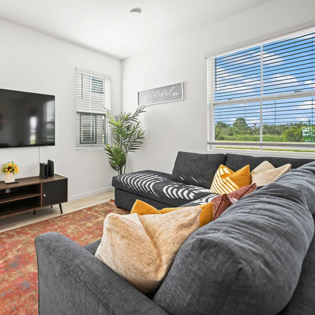 A modern living room features a large TV mounted on a white wall, a dark sectional sofa with colorful cushions, and a small table with a plant and a vase of flowers.