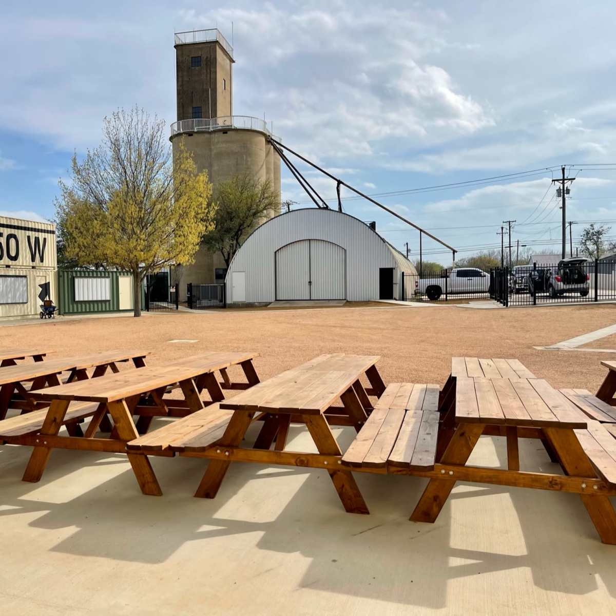 The scene shows a row of wooden picnic tables in an outdoor area with a large industrial building and a tower in the background under a partly cloudy sky.