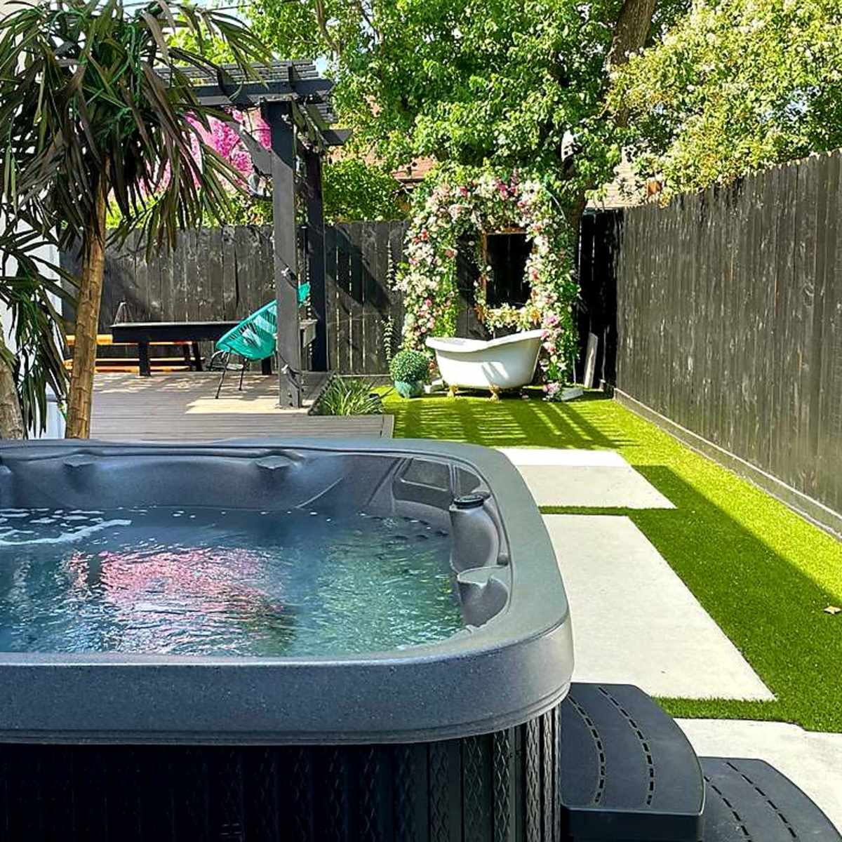 A hot tub sits on a patio surrounded by greenery and decorative elements, including a bathtub and artificial grass.