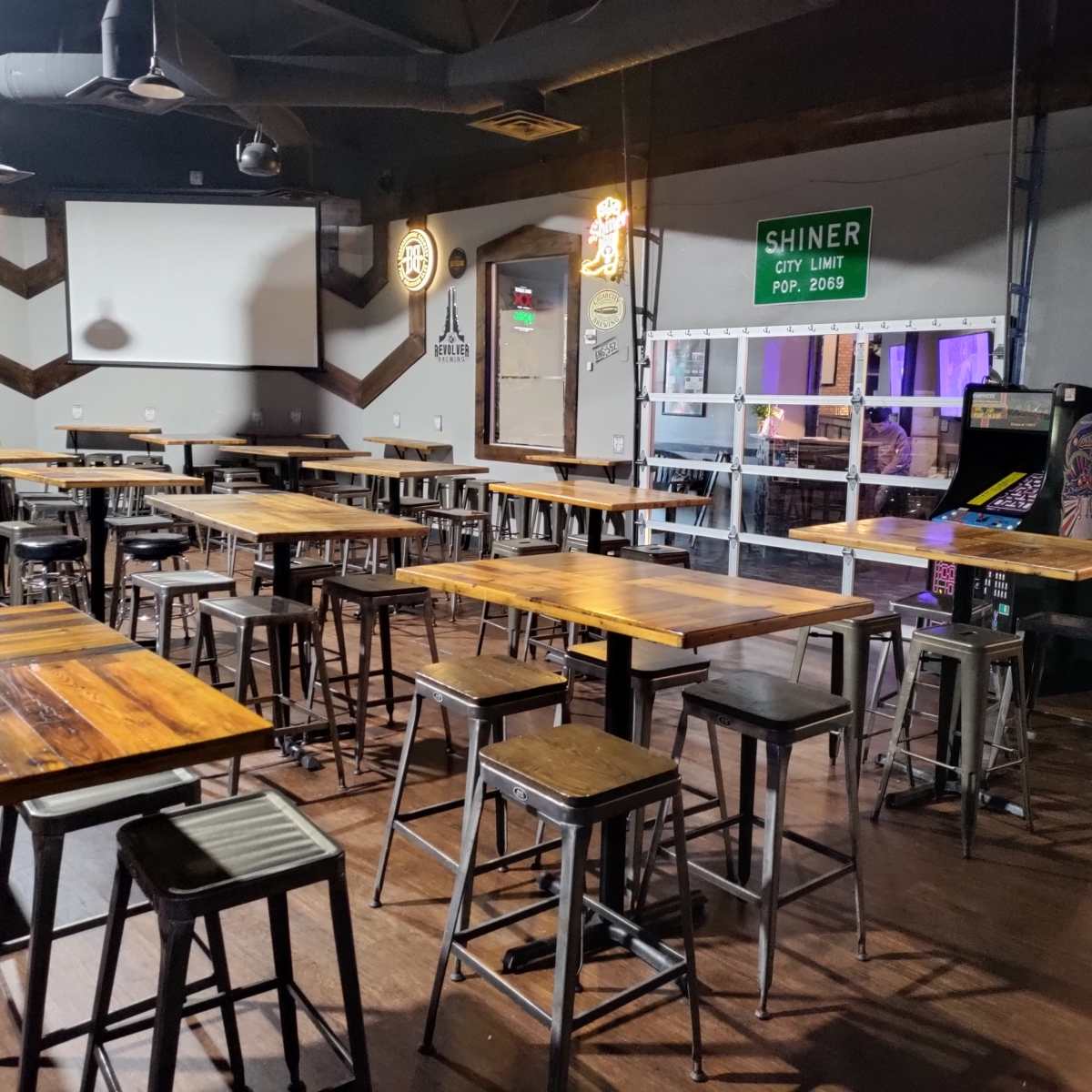 An empty dining area with wooden tables and metal stools, featuring a bar arcade machine in the corner and a large screen on the wall.