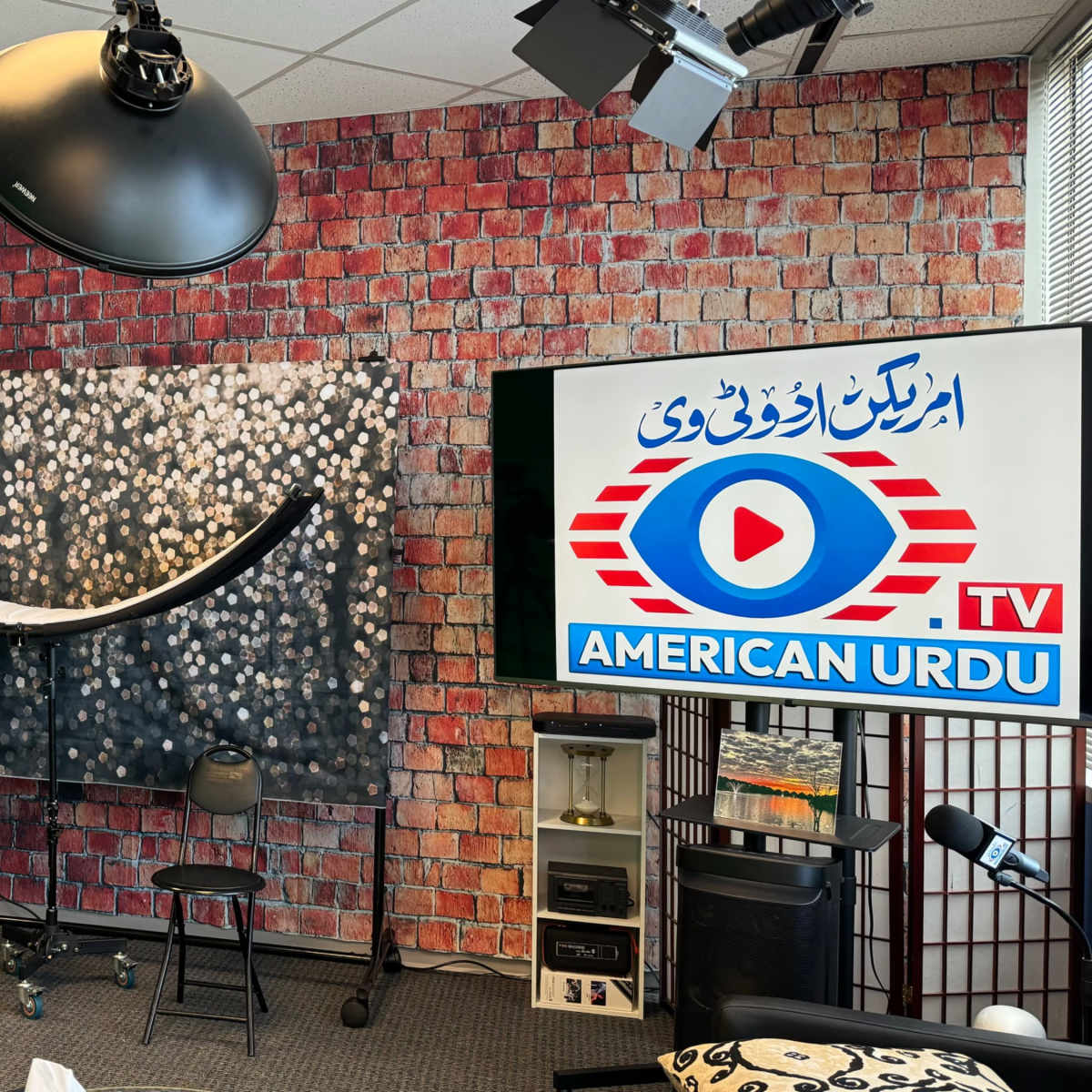 A studio setup with photography equipment, including softbox lights and a backdrop, alongside a television displaying a logo for "American Urdu TV."