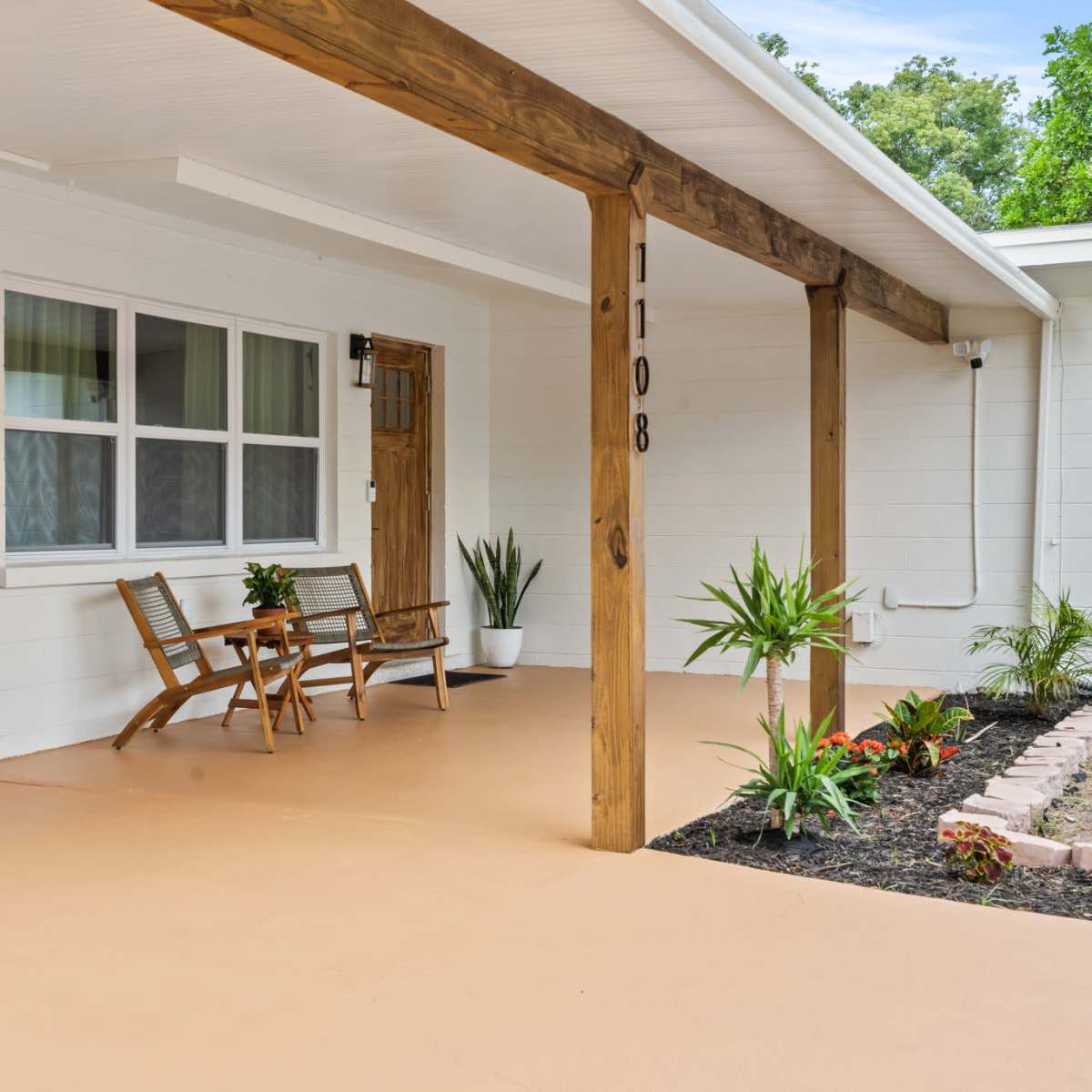 A porch area with a wooden ceiling and support beams, featuring two chairs and a small table on a concrete floor, alongside decorative plants and a well-maintained garden border.