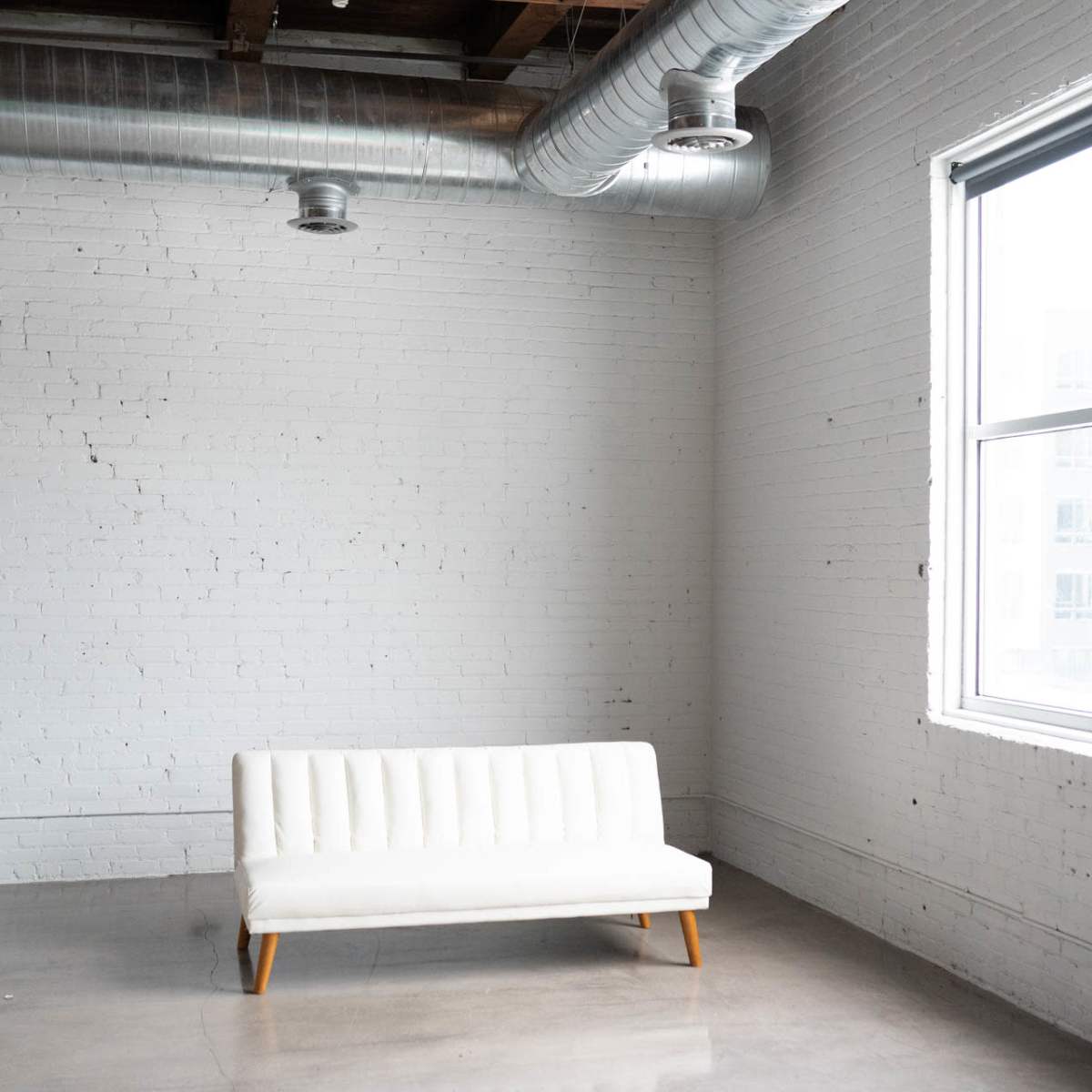 A white sofa is positioned against a light gray brick wall in a spacious, minimally decorated room with large windows and exposed ductwork.