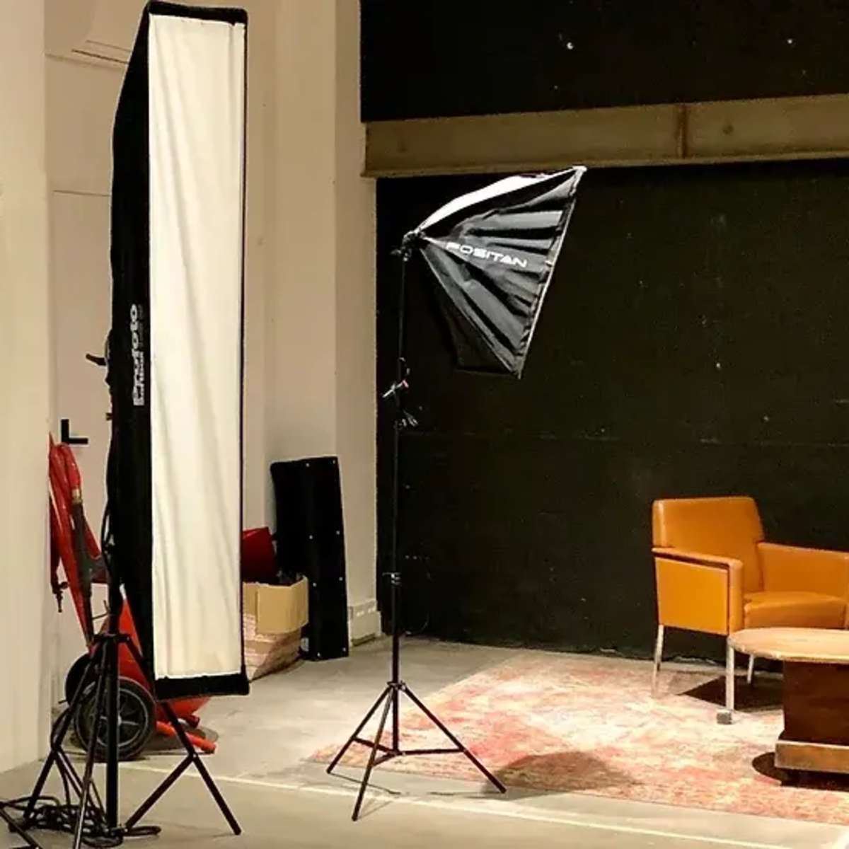 A photography studio setup features two orange chairs facing a coffee table, surrounded by lighting equipment and backdrop drapes.