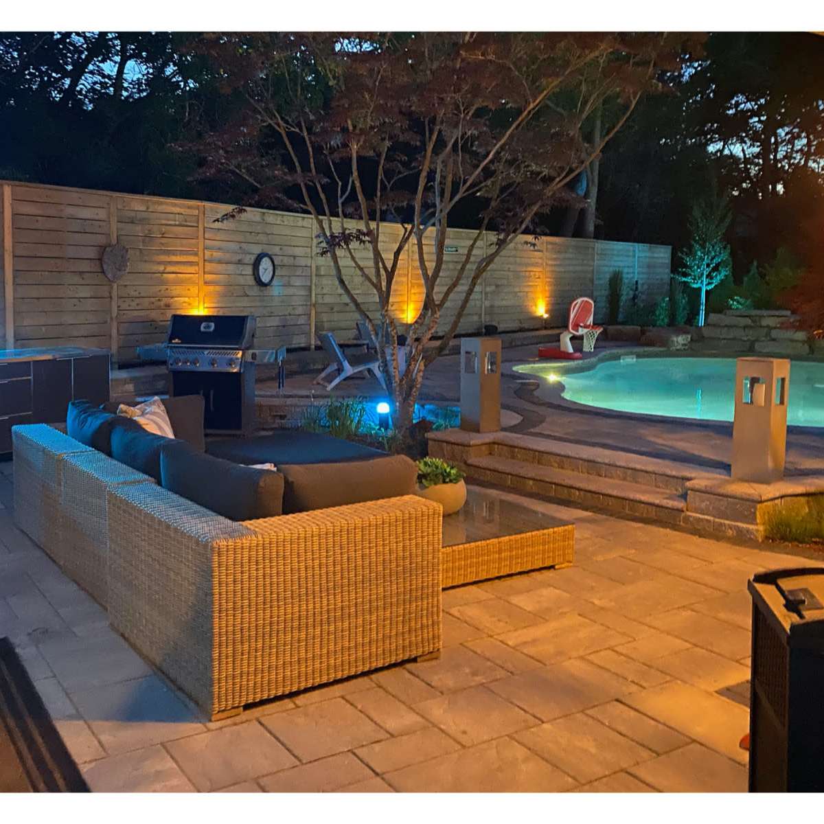A patio area with a sectional sofa, a grill, a pool, and ambient lighting in a landscaped backyard.