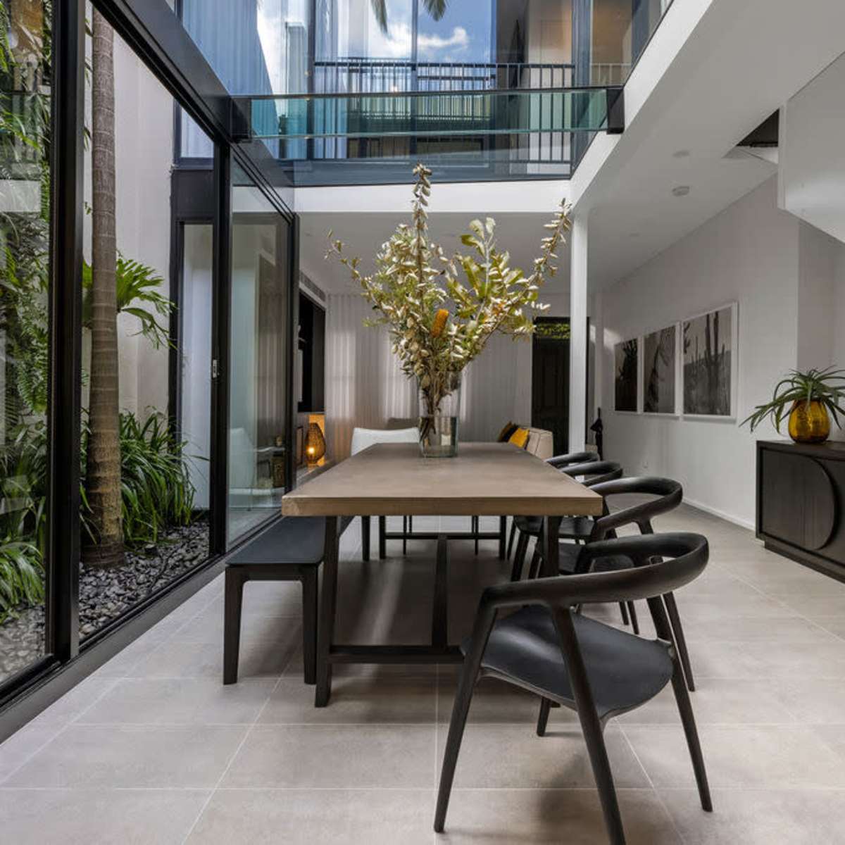 A modern dining area with a long wooden table, black chairs, and large windows that overlook a green outdoor space.