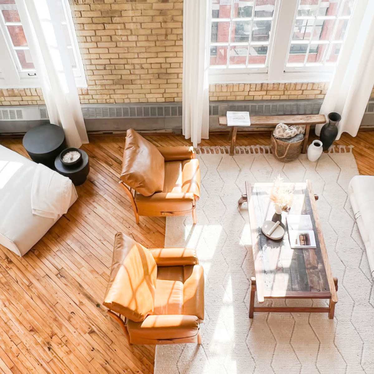 A bright living room with wooden floors, large windows, and a mix of furniture including two armchairs, a white sofa, and a coffee table.