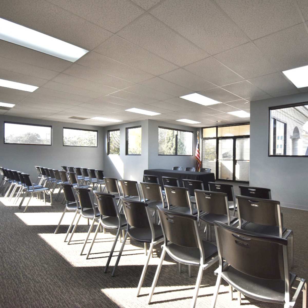 A vacant meeting room features rows of black chairs facing a front area with large windows and a door leading outside.