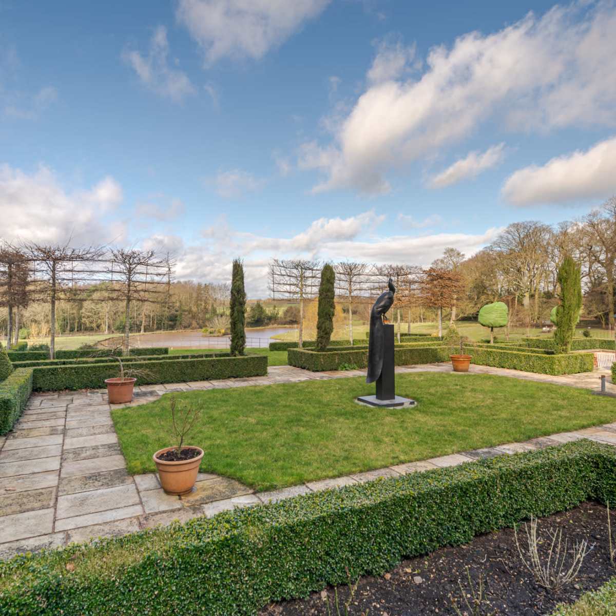A neatly maintained garden features a central statue surrounded by manicured hedges and potted plants under a partly cloudy sky.