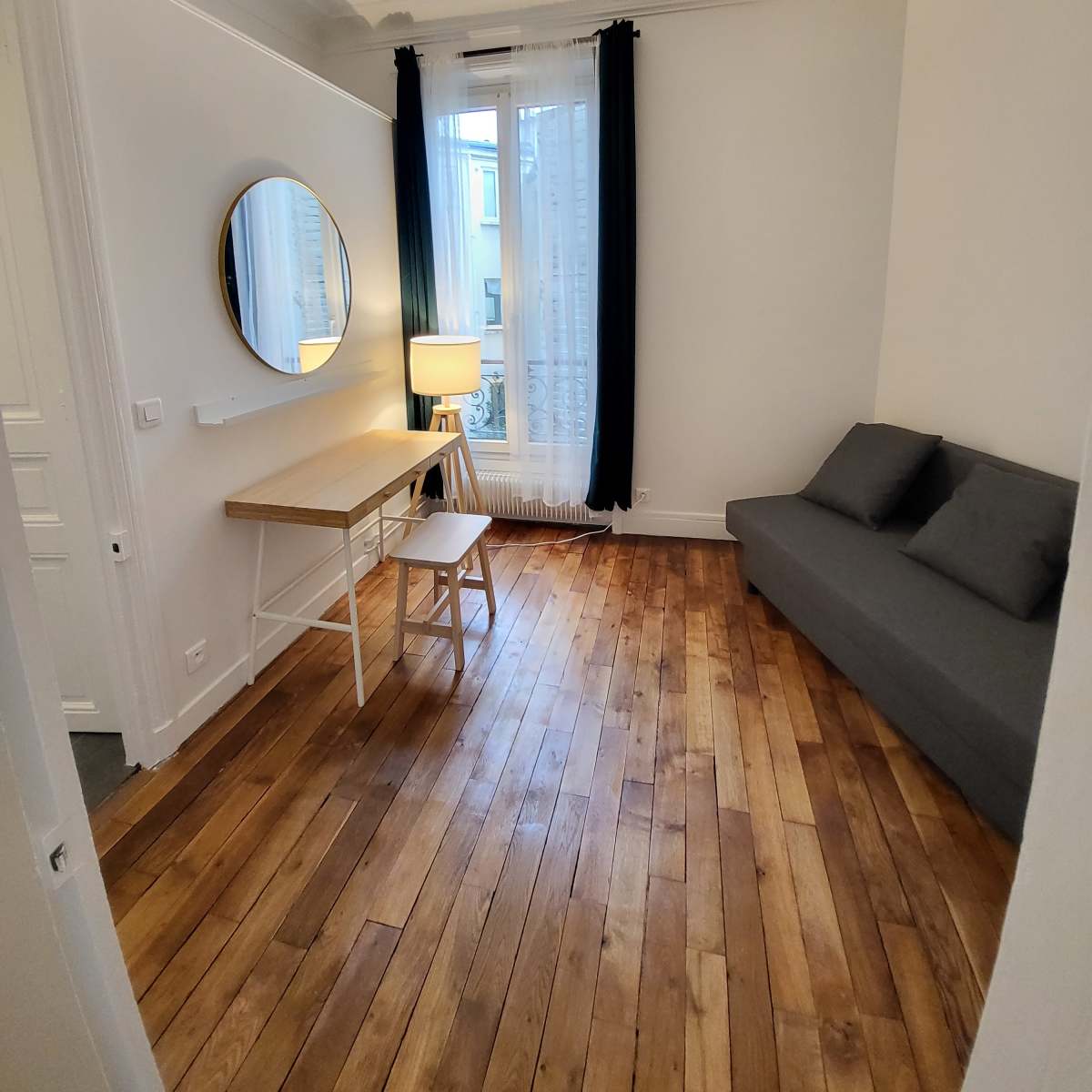 A small, well-lit room featuring a desk with a chair, a round mirror on the wall, a sofa, and a lamp, all set on wooden flooring.