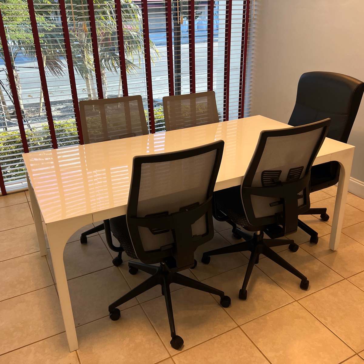 A white conference table with five chairs is positioned in a room with blinds and a view of outdoor greenery.