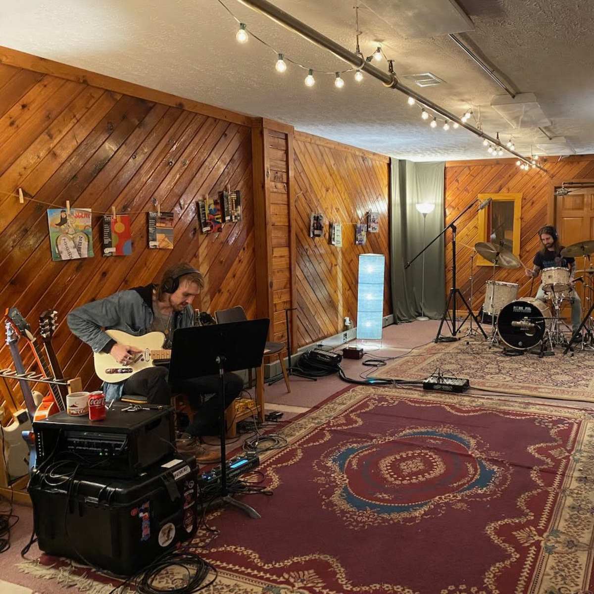 Two musicians are recording in a wood-paneled room, with one playing an electric guitar and the other on drums.