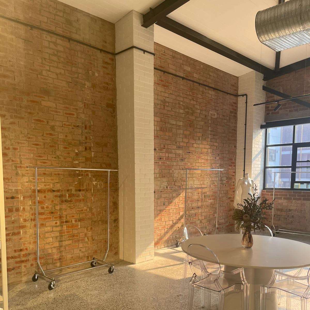 A spacious room with brick walls, a round white table, ghost chairs, and clothing racks on wheels.
