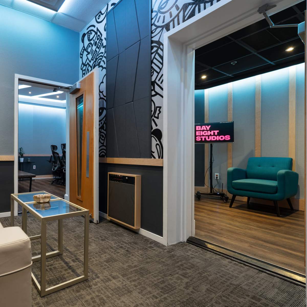A modern studio lounge area with a white couch, a glass coffee table, and a doorway leading to a soundproof recording room featuring teal furniture and backlit studio signage.