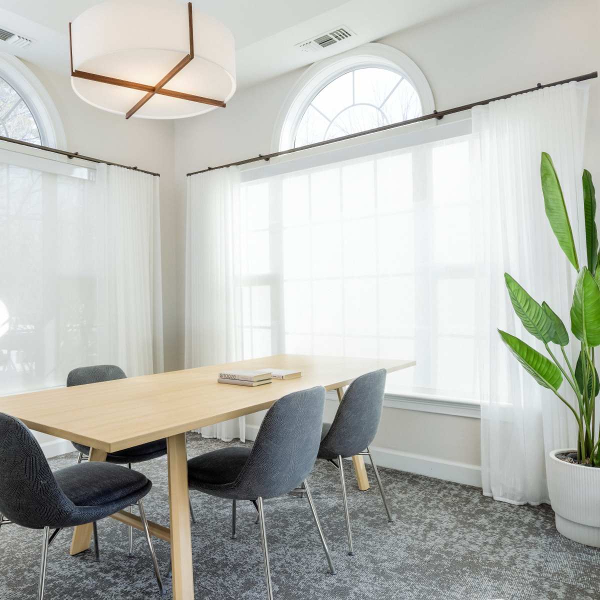 A wooden dining table is surrounded by four gray chairs in a brightly lit room with large windows and sheer curtains.