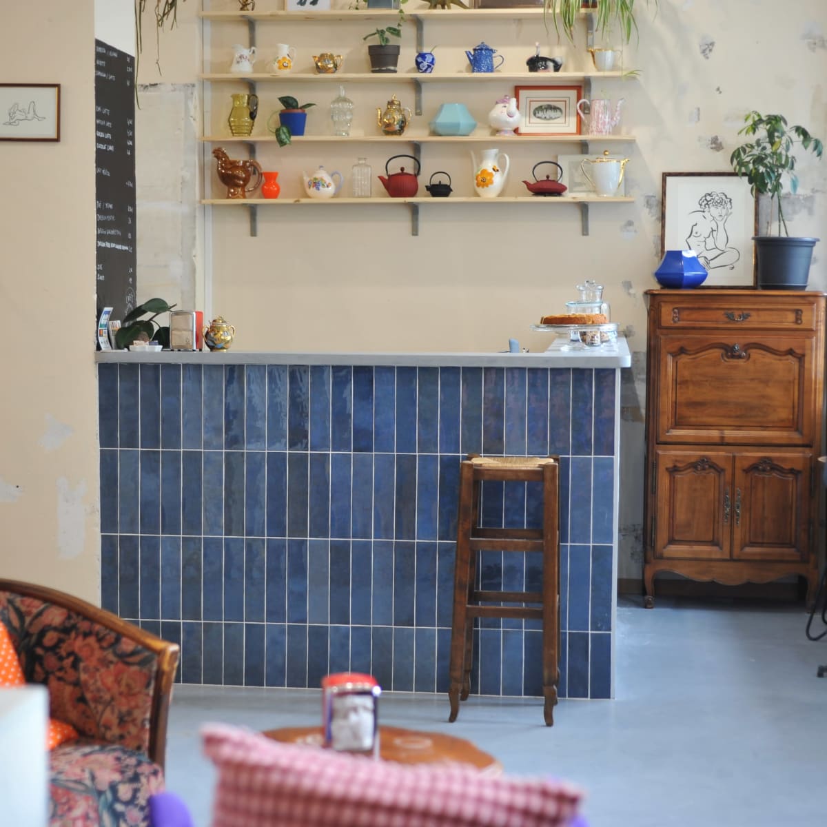 A cozy cafe features a blue tiled counter, shelves adorned with teapots, and wooden furniture including a vintage cabinet and a small table.