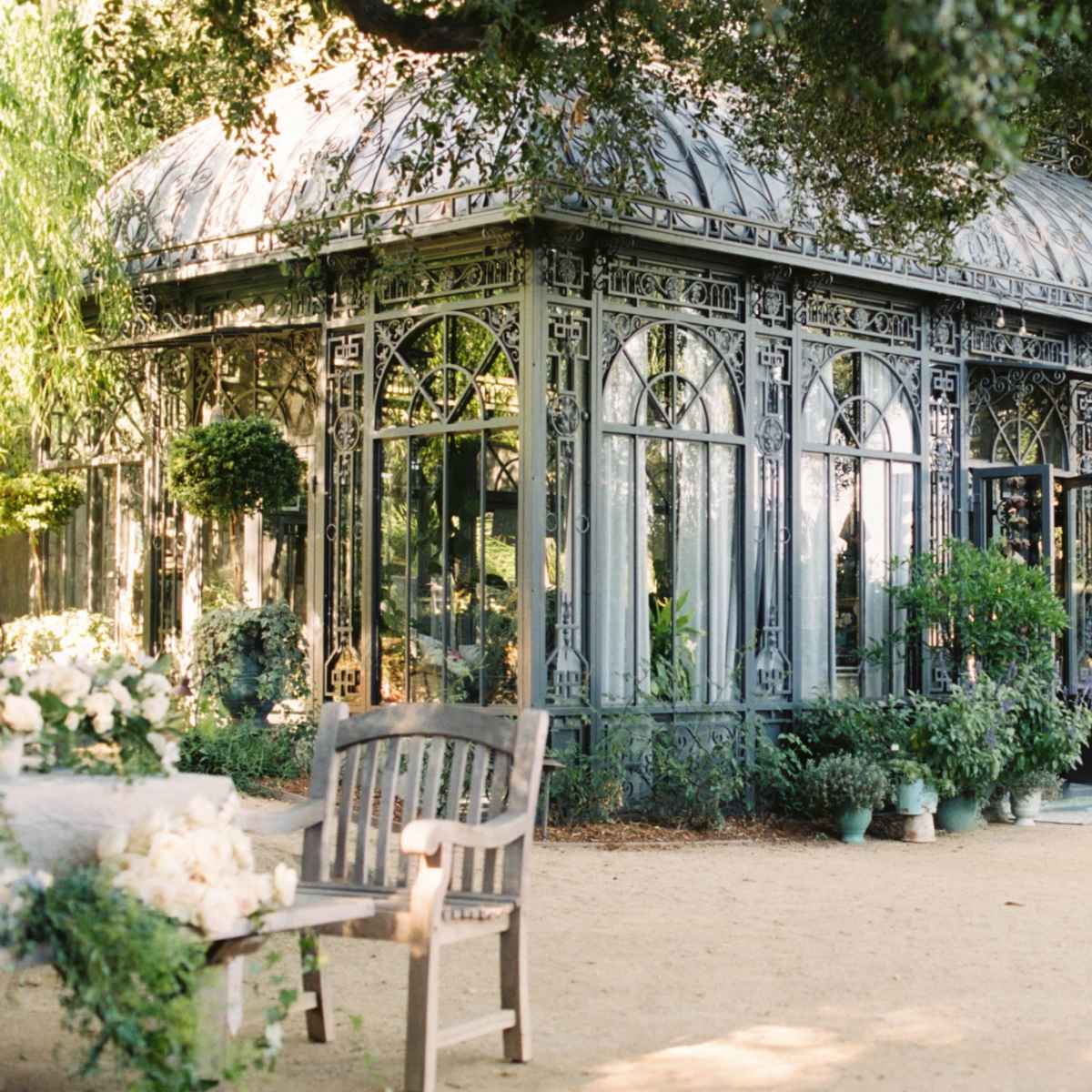 A glass conservatory with intricate metalwork is surrounded by greenery and potted plants in a garden setting.