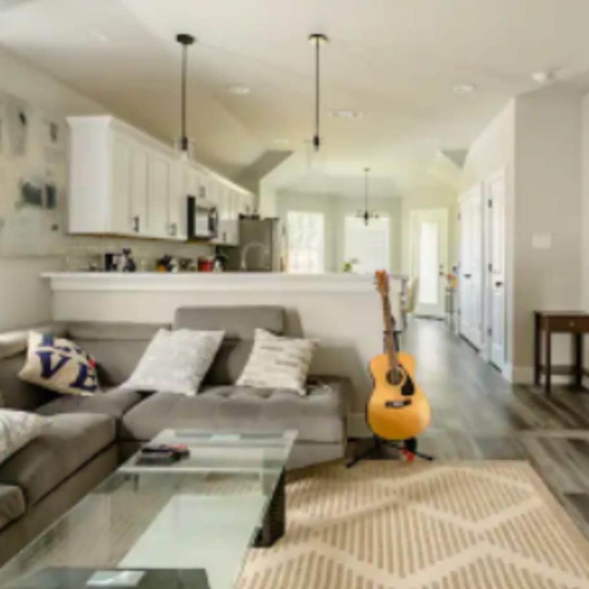 A living room features a gray sectional sofa, a coffee table, an acoustic guitar standing against the wall, and a view into a kitchen area and hallway.