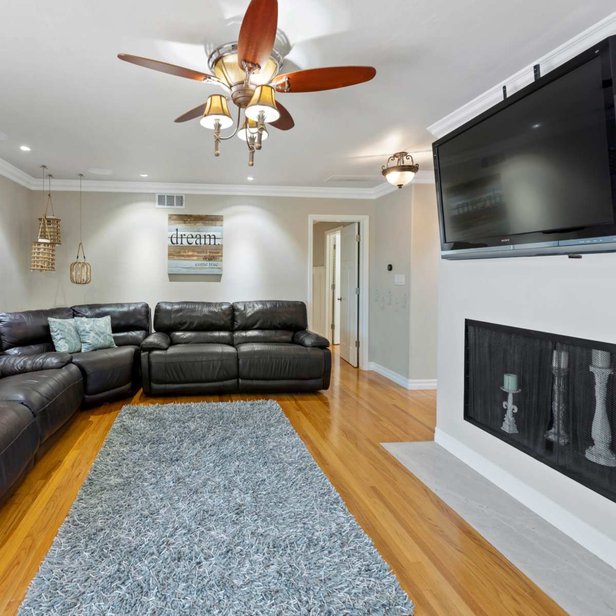 The living room features a large leather sectional sofa, a flat-screen TV mounted on the wall, and a decorative area rug on polished hardwood floors.