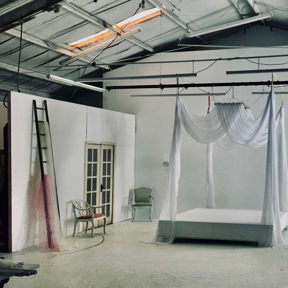 A spacious, empty studio features a white bed draped with sheer fabric, accompanied by vintage furniture and soft lighting.
