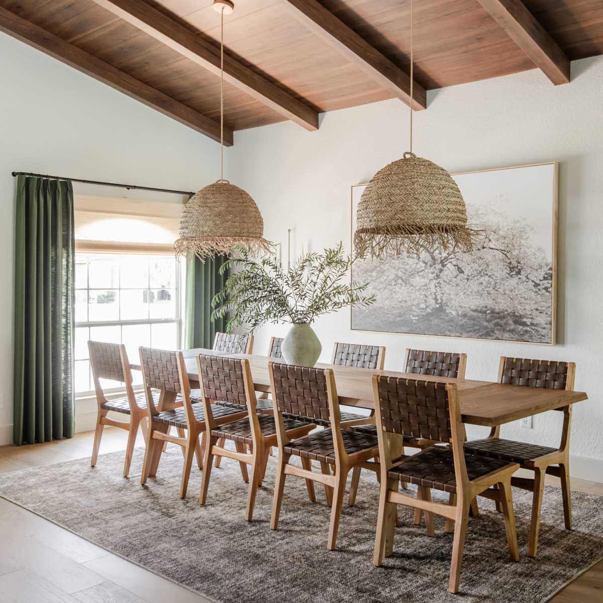 A spacious dining room features a long wooden table surrounded by eight woven chairs, with large pendant lights hanging above and a framed artwork on the wall.