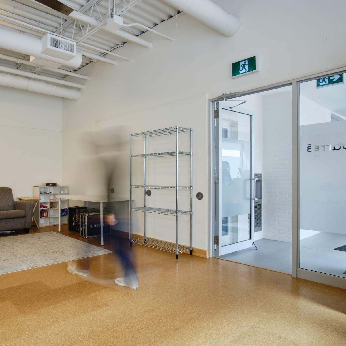 A person walks through a minimalist waiting area with a couch, shelving unit, and an exit door.