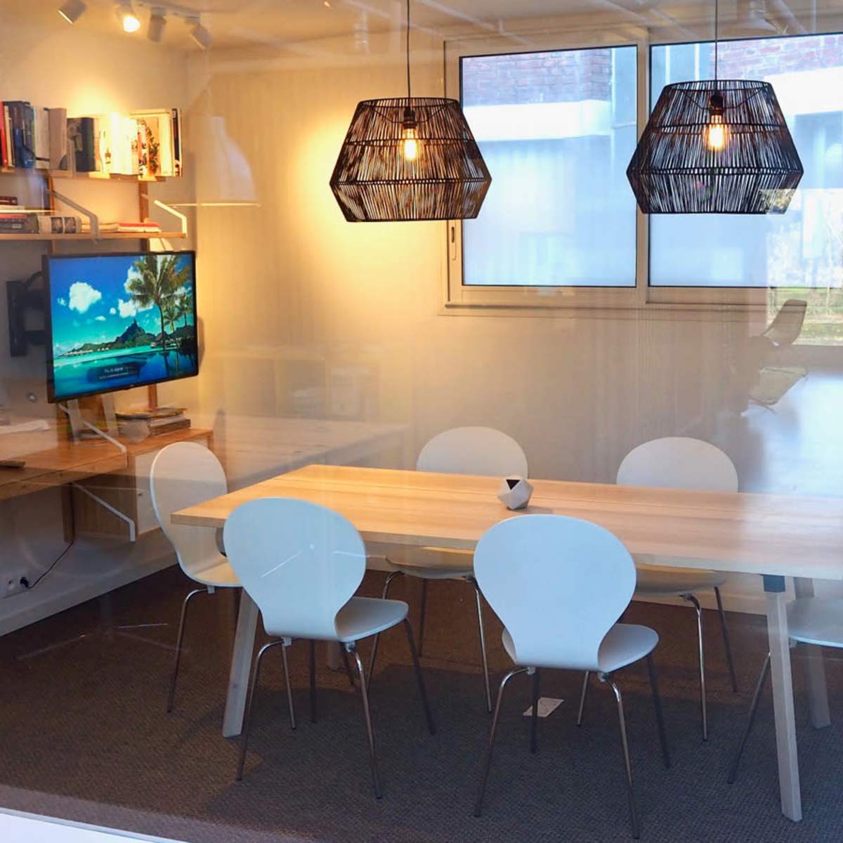 A modern conference room featuring a wooden table surrounded by white chairs, a wall-mounted television, and two pendant lights above the table.