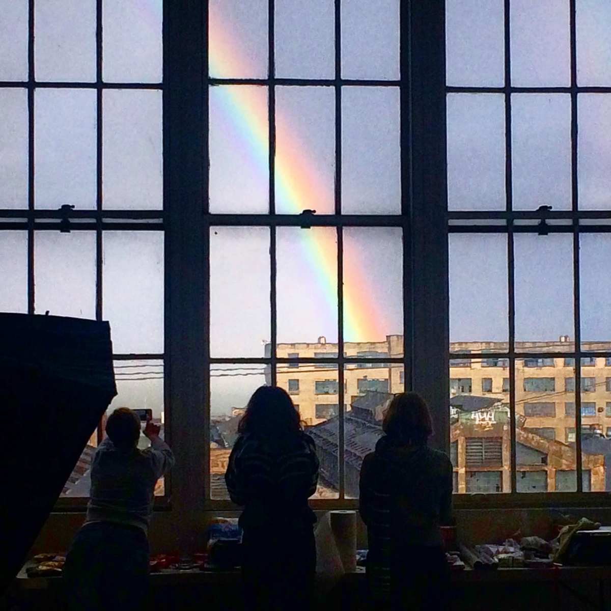 Three people stand by large windows, observing a rainbow arching over an industrial building.